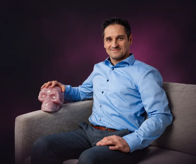 Alex sitzt lächelnd auf einem Sofa, die Hand auf einem Rosenquarz-Schädel
